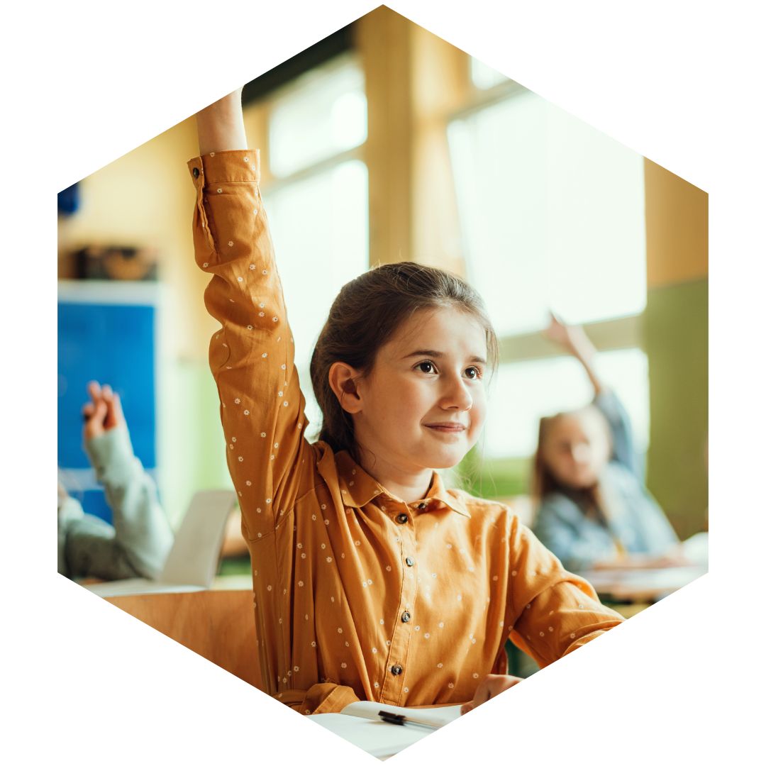 Young girl seated at a desk, raising her hand in a classroom.