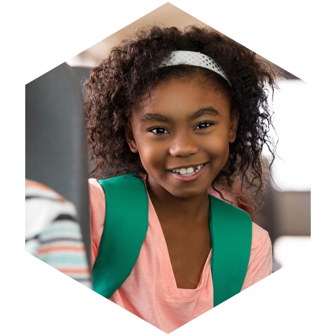 Smiling young girl with a headband and backpack looking out from a bus seat.