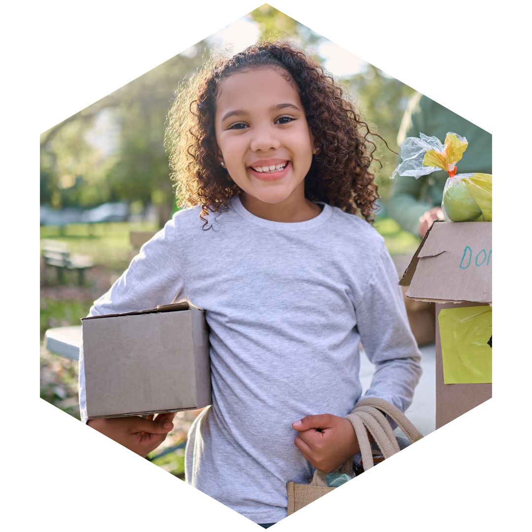 Smiling young girl holding a box and bags with a large box of donations behind her.