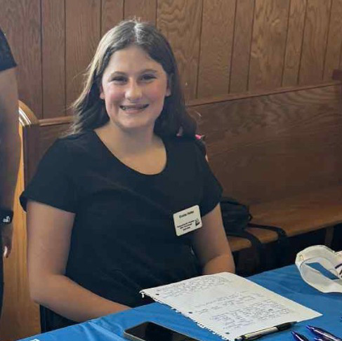 Smiling teenage girl seated at a table with a handwritten paper in front of her.