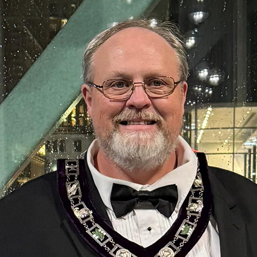 Smiling man with short hair, a beard, and glasses in Masonic dress standing indoors.