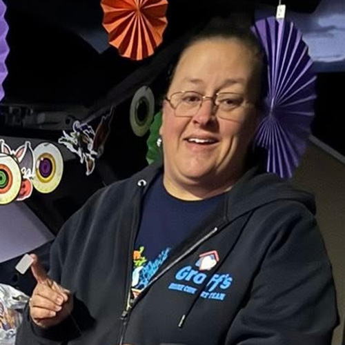 Smiling woman with glasses and her hair pulled back standing in front of colorful Halloween decorations.