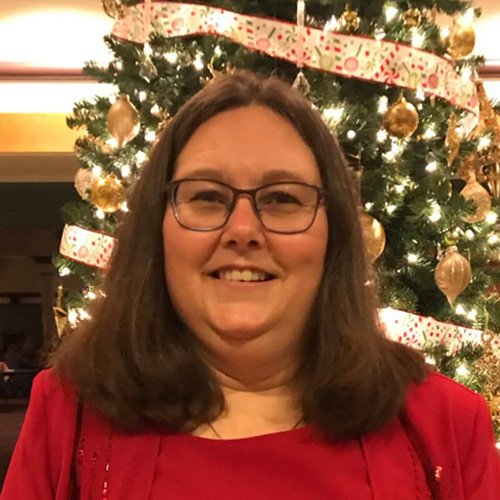Smiling woman with glasses and brown hair standing in front of a decorated Christmas tree.