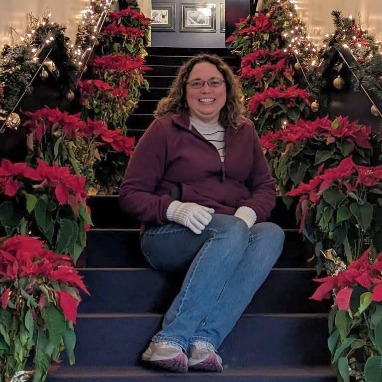 Smiling woman in glasses, coat and gloves seated on a poinsettia-lined staircase.