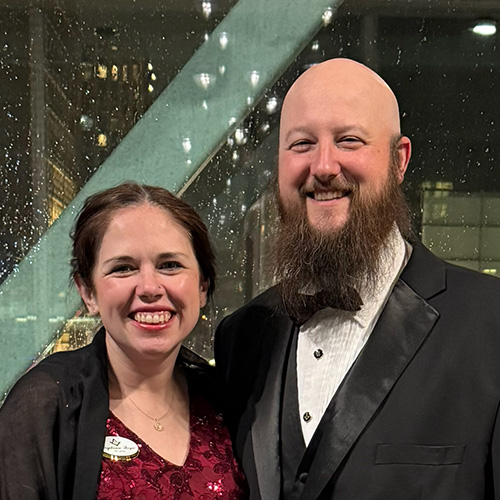 Smiling couple dressed in formal attire, standing indoors in front of a window.