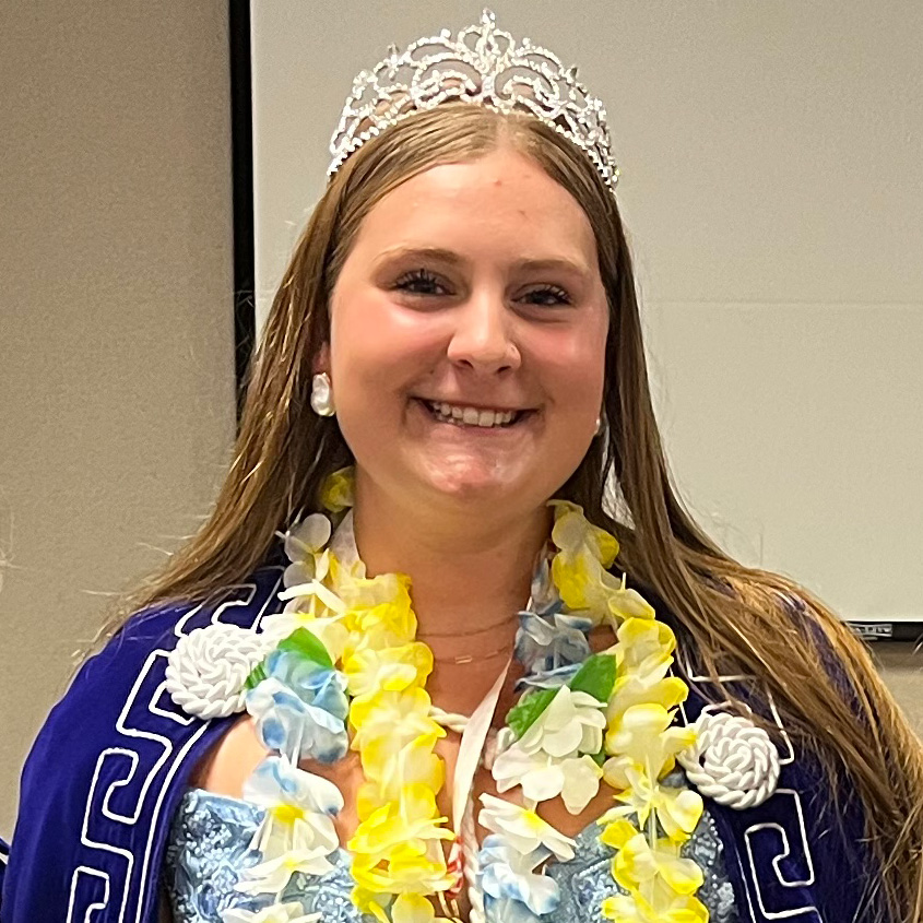 Smiling young lady wearing a crown, a blue cape and floral leis while standing indoors.