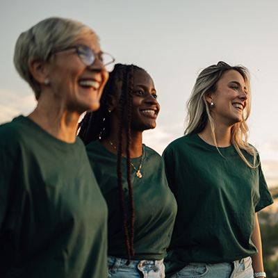 Three smiling adult women wearing green shirts and standing outdoors.