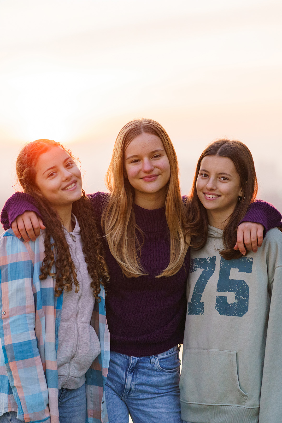 Three smiling girls with arms around each other, the sun is setting behind them.