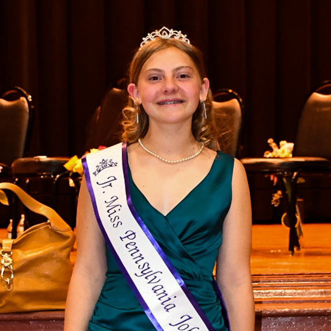 Smiling girl with a crown, sash and green dress standing in front of a stage.