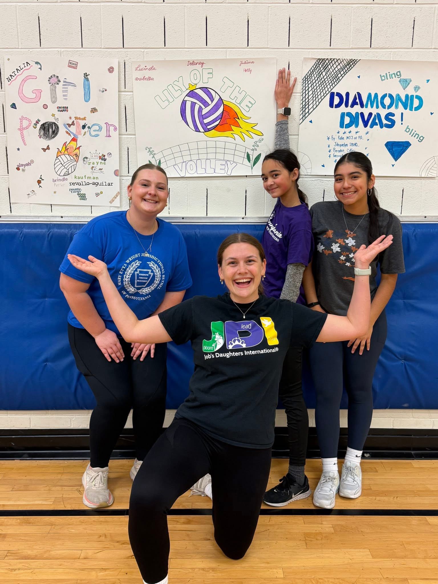 Group of smiling girls posed together in a gym with volleyball signs behind them.