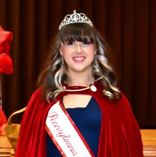 Smiling girl in a red cape and crown standing in front of a stage.