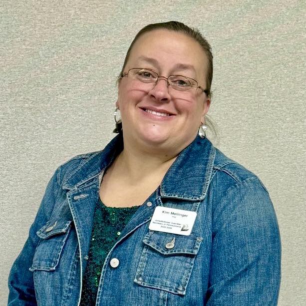 Kim Mellinger Smiling woman with glasses wearing a jean jacket in front of a cream-color walled.